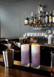 Two tall glasses filled with a layered purple and white drink, garnished with lemon twists, are placed on a bar counter. A metal shaker sits nearby. In the background, glasses and bottles are neatly arranged on shelves.