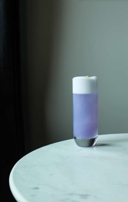 A tall glass filled with a lavender-colored drink topped with white foam and a lemon peel garnish sits on a round, white marble table against a dark background.