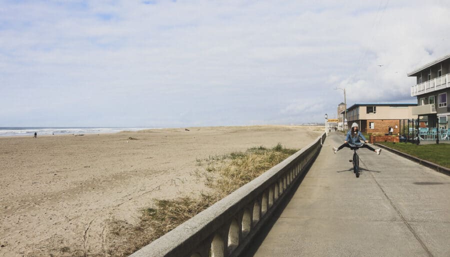 A person rides a bicycle with legs outstretched on a paved walkway next to a sandy beach. The sky is mostly cloudy, and a few buildings line the path on the right side of the image.