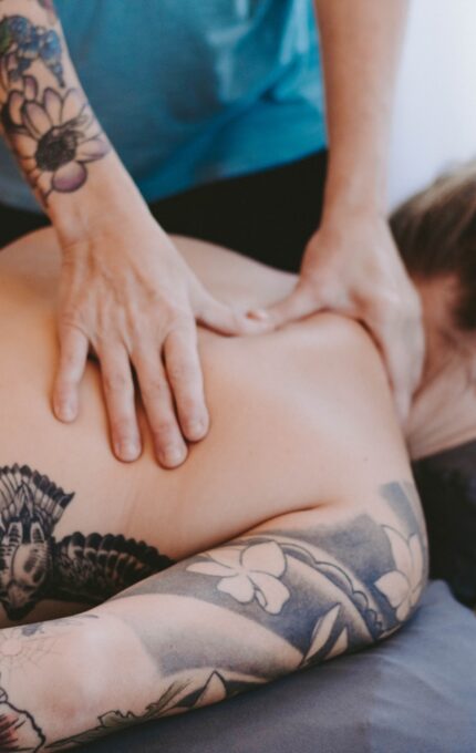 A person with tattoos on their arm and back lies face down on a massage table while another person massages their shoulders and upper back.