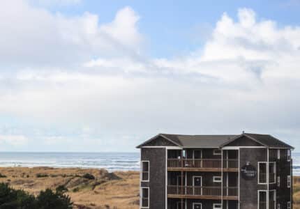 A coastal building with multiple balconies stands near grassy dunes, overlooking a vast ocean under a partly cloudy sky. The structure is three stories high and wears wooden siding. Waves can be seen in the distance.