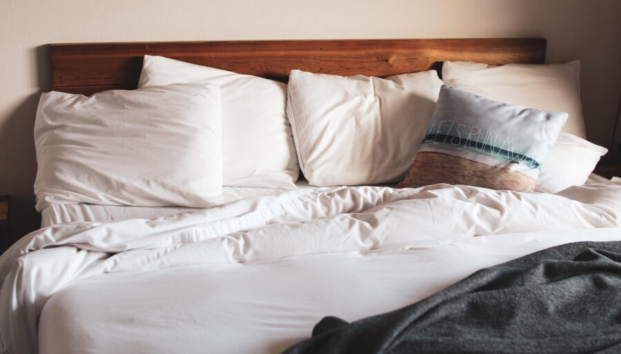 A bed with white sheets and pillows, including a decorative pillow with a landscape design and text, against a wooden headboard. A dark blanket is partially visible at the bottom of the bed.