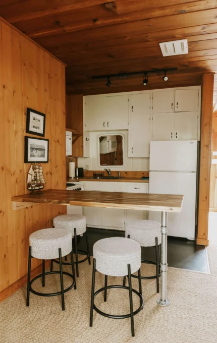 Cosy hotel kitchenette with wooden decor, high-top table, white stools, and modern appliances for a relaxing stay.