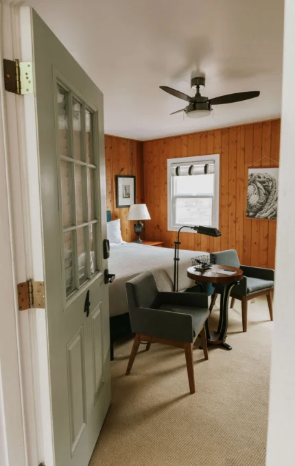 Cosy hotel room with wooden walls, double bed, modern lamp, and armchairs, offering a warm, relaxing stay.