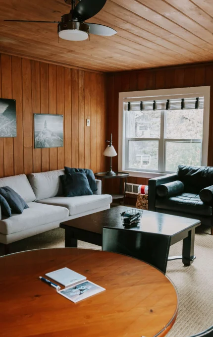 Cosy hotel lounge with wooden walls, modern sofa, armchair, large windows, and round table for a relaxing stay.