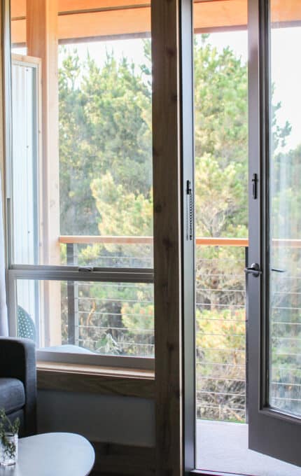 A cozy living room with a gray sofa and assorted cushions, next to a round coffee table with a small vase. Large glass doors open to a wooden balcony, revealing a view of dense green trees. White curtains hang by the windows.