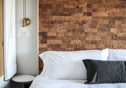 A neatly made bed with white pillows and a dark gray cushion is positioned against a textured brown wall. A small round white side table is nearby, holding a modern light fixture with a clear glass shade hanging above.