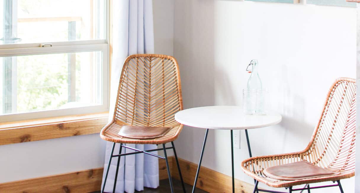 A small seating area with two wicker chairs and a round white table. There's a glass bottle and two glasses on the table. A window with sheer curtains is on the left, and framed artwork is partially visible on the wall.