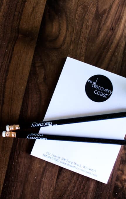 Two black pencils and a notepad with "Discovery Coast" branding sit on a wooden table beside a small potted plant with green leaves.