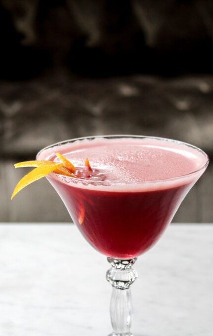 A martini glass filled with a bright red cocktail, garnished with a twist of orange peel, is placed on a white marble surface. The background is softly blurred, showcasing a dark, plush texture.