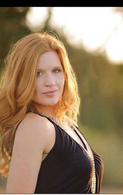 A person with long red hair wearing a sleeveless black dress stands outdoors. The background is a blurred natural setting with green foliage, suggesting a warm, sunny day.