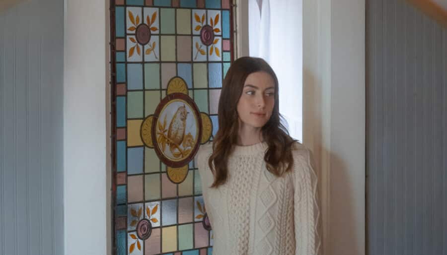 A woman wearing a cream knit sweater stands indoors, her reflection seen in a hexagonal mirror. Behind her is a door and a decorative stained glass window with floral and geometric patterns.