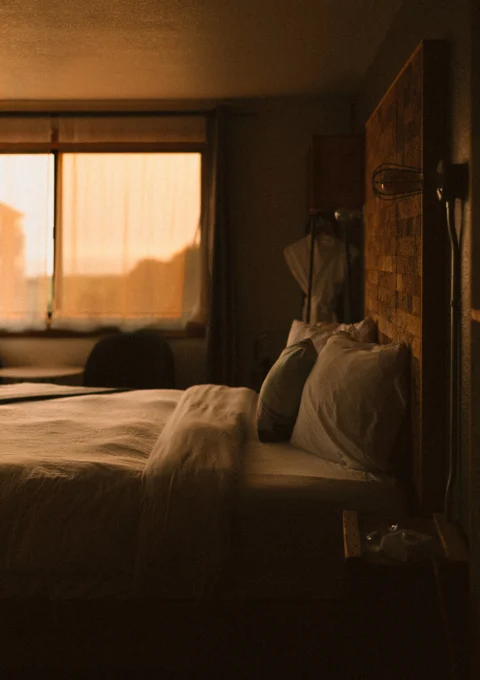 Cosy king bed with plush pillows, textured wooden headboard and warm sunrise light through sheer curtains, peaceful boutique hotel room