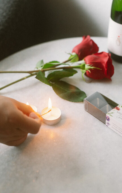 A person lights a small white tealight candle on a round, light-colored table. Two red roses, a matchbox, and a wine bottle are also on the table.