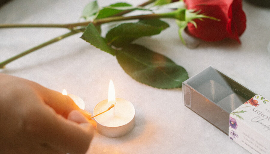 A person lights a small white tealight candle on a round, light-colored table. Two red roses, a matchbox, and a wine bottle are also on the table.