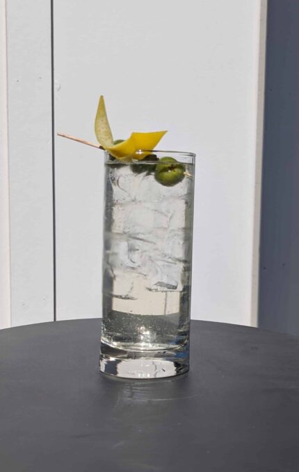 A tall glass filled with a clear beverage and ice cubes, garnished with a green olive and a lemon twist on a wooden skewer, sits on a dark surface against a light-colored background.