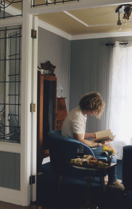 A person with curly hair sits in a blue chair by a window, reading a book in a softly lit room. A small table with food and flowers is nearby. The adjacent bathroom with a bathtub is visible through an open door.