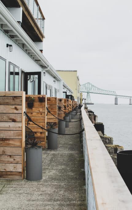 The image shows a row of waterfront buildings with wooden balconies, facing a large body of water. A distant bridge extends across the water under a cloudy sky. The scene is calm and monochromatic.