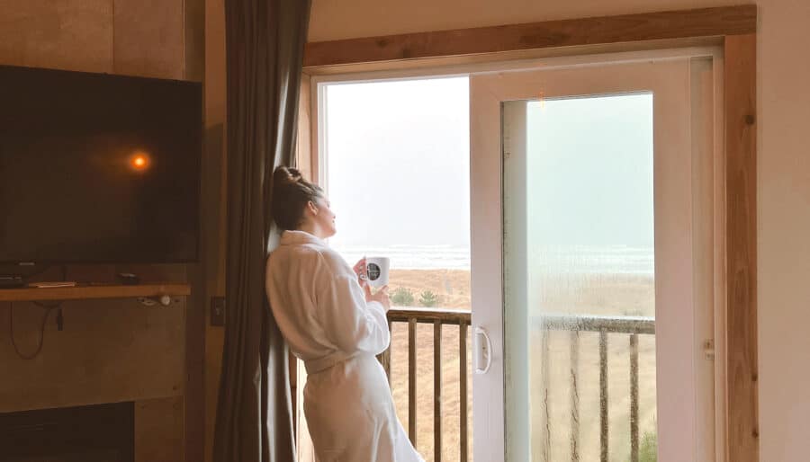 A person in a white bathrobe stands by a sliding glass door, holding a cup and gazing outside. The room has a wall-mounted TV, a fireplace, and wooden flooring. The view outside shows a grassy field and misty sky.
