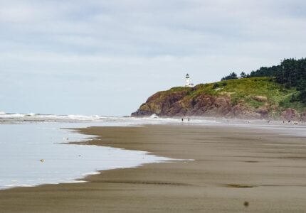 A scenic view of a sandy beach with gentle waves rolling in. In the distance, a rocky cliff is topped with a lighthouse. The shoreline is lined with trees, and a few people can be seen walking along the water's edge.