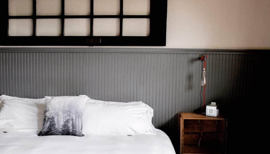 A minimalist bedroom features a low bed with white sheets and a decorative pillow. A wooden crate serves as a nightstand, holding a small device. Above, a window with a grid pattern adds contrast to the room's neutral tones.