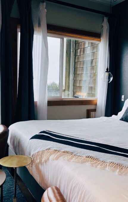 A modern bedroom with a large bed featuring a dark blanket and patterned pillows. The room includes a leather chair, a small side table, and large windows with sheer curtains, offering a view of a wooden balcony and greenery outside.