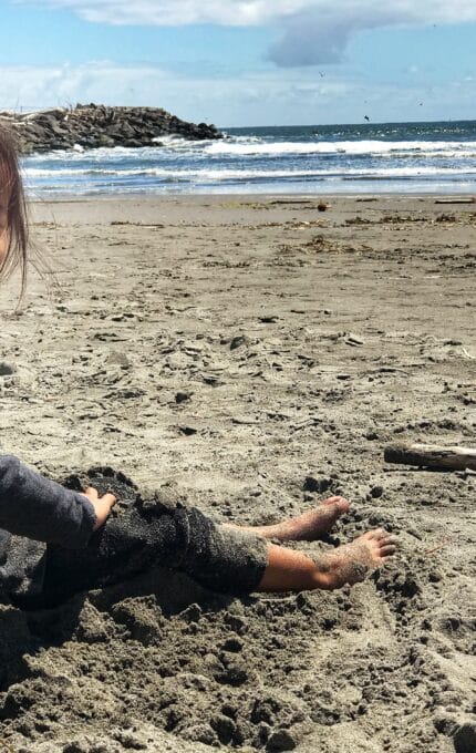 A young child sits on a sandy beach, smiling and looking at the camera. The ocean waves are visible in the background, under a partly cloudy sky. The child is dressed in a dark long-sleeved outfit, with bare feet partially buried in the sand.