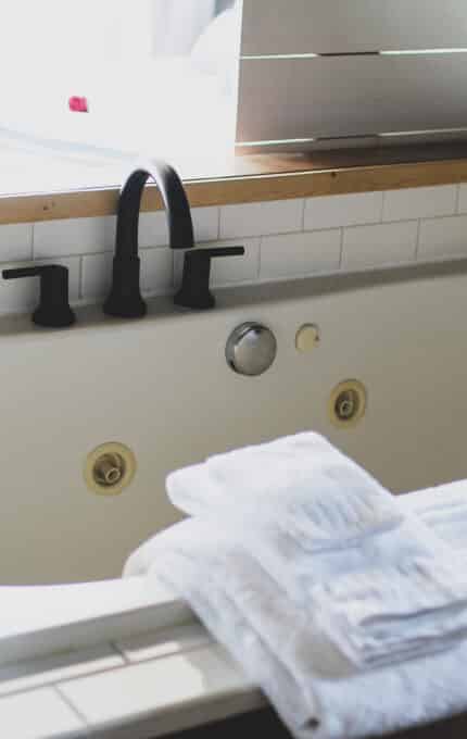 A bathroom scene featuring a white bathtub with black fixtures and a wooden frame. Several towels are neatly folded on the edge of the tub, and a small yellow rubber duck is placed on one of the towels.