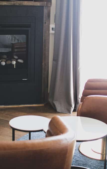 The image shows a cozy living room with two brown leather chairs facing a modern fireplace. A small round table is positioned between the chairs. The room has wooden flooring and a large window with a curtain partially drawn to the side.