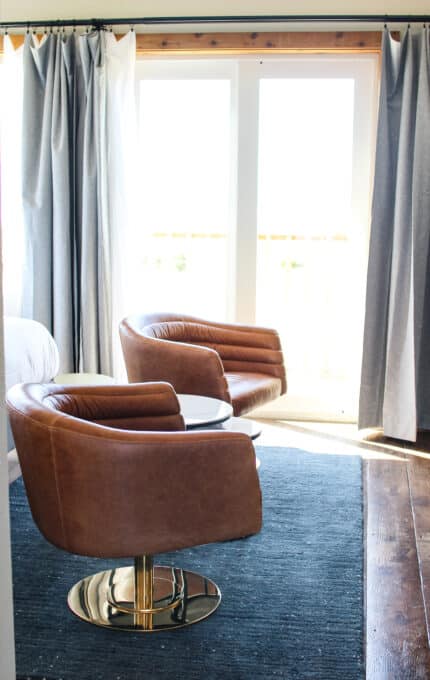 Two brown leather swivel chairs sit on a dark rug in front of a round white table. The area is well-lit by natural light coming through large glass doors, partially covered by gray curtains. The wooden floor adds a contrasting texture.