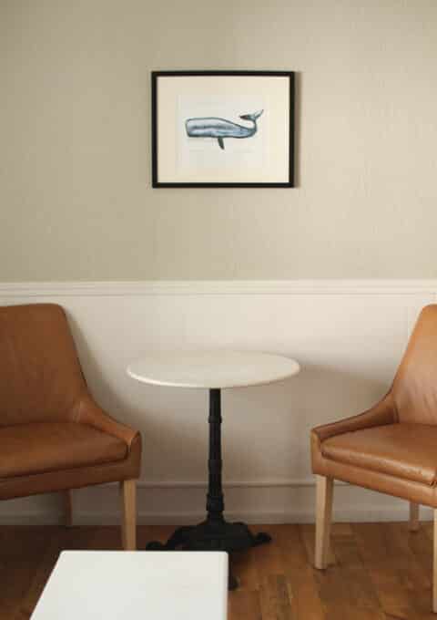 Two brown leather chairs flank a small round white table with a black pedestal base. A framed illustration of a blue whale hangs on a cream-colored wall above the table. The floor is wooden.