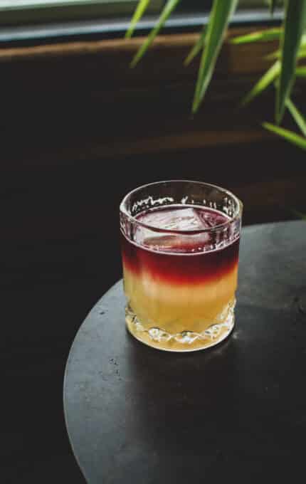 A layered cocktail in a glass with a purple top layer and a yellow bottom layer sits on a dark round table. In the background, green leafy plant details are visible.