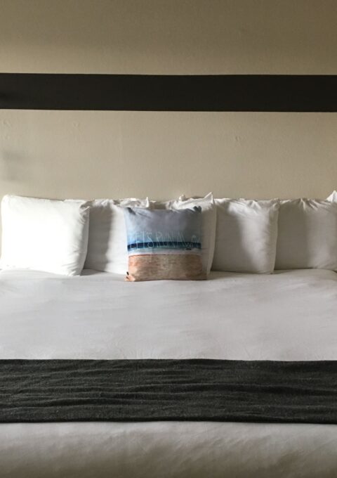 A neatly made bed with four white pillows, one decorative pillow with text, and a gray blanket across the foot of the bed. Two red cords with light bulbs hang from a black horizontal board above the headboard. The wall is beige.