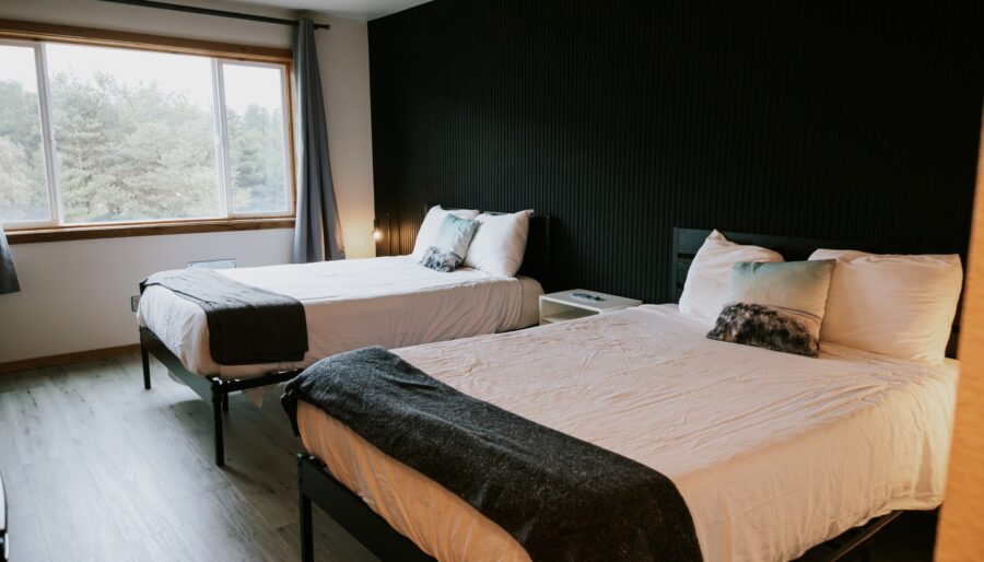 A modern bedroom with two double beds, each with white bedding and dark gray throws, against a black accent wall. A large window lets in natural light, and a nightstand sits between the beds.