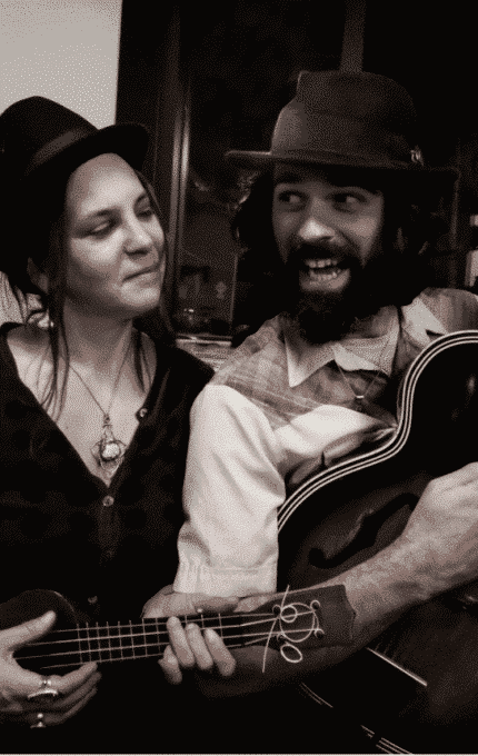 A man and woman are smiling at each other while holding musical instruments. The man has a guitar, a hat, and a beard. The woman has a ukulele and is also wearing a hat. They are indoors with a dark window behind them.