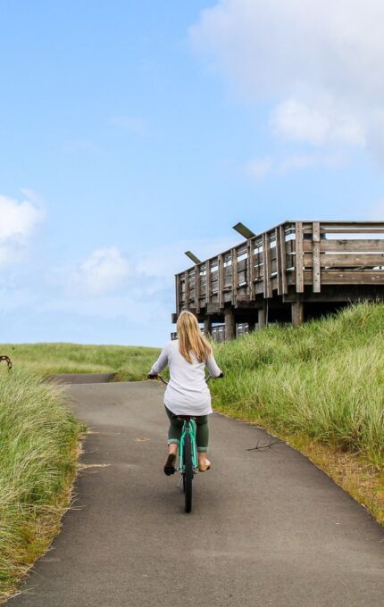 A person with long blonde hair rides a bicycle on a paved path through grassy dunes under a partly cloudy sky. A wooden observation deck stands on stilts to the right of the path.