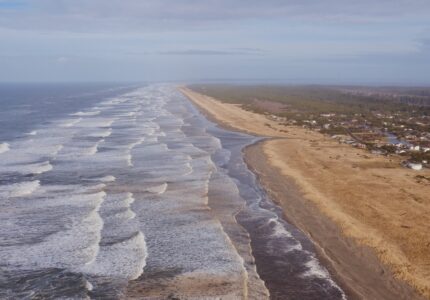 An aerial view of a coastline featuring a long sandy beach adjacent to an ocean with gentle waves. On one side of the beach are small buildings and houses, with a forested area further inland. The sky is partly cloudy.