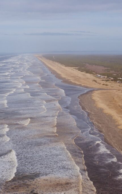 An aerial view of a coastline featuring a long sandy beach adjacent to an ocean with gentle waves. On one side of the beach are small buildings and houses, with a forested area further inland. The sky is partly cloudy.