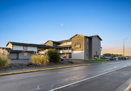 A three-story modern building with a sign reading "Adrift" in front, situated beside a paved road with parked cars. The building has large windows and balconies. Landscaping around the building includes grasses and bushes. The sky is clear with a crescent moon visible.