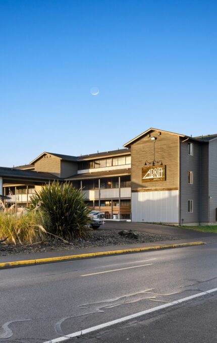 A three-story modern building with a sign reading "Adrift" in front, situated beside a paved road with parked cars. The building has large windows and balconies. Landscaping around the building includes grasses and bushes. The sky is clear with a crescent moon visible.