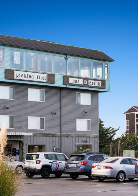 A multi-story building houses the Adrift Hotel, with the Pickled Fish eat & drink restaurant on the top floor. Several cars are parked in the adjacent lot, and another building is visible in the background. Ornamental grass is in the foreground.