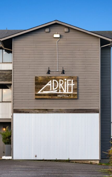 The exterior of Adrift Hotel, a three-story building with a wooden facade. The hotel sign is prominently displayed in the center, and there is a blue door and a bench on the ground level. There are some grasses and a clear blue sky in the background.