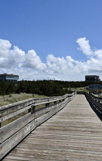 A wooden boardwalk leads through grassy dunes toward several distant buildings under a partly cloudy sky. Dense shrubbery lines the path, and rolling hills are visible in the background.