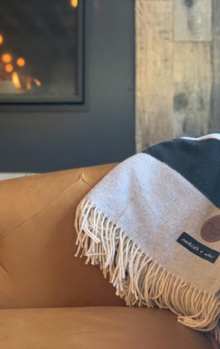 A cozy, brown leather chair is positioned near a modern fireplace. A patterned blanket with fringe detailing is draped over the arm of the chair. The background features rustic wooden paneling and the fireplace emits a warm and inviting glow.