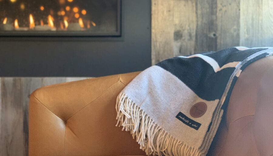 A cozy, brown leather chair is positioned near a modern fireplace. A patterned blanket with fringe detailing is draped over the arm of the chair. The background features rustic wooden paneling and the fireplace emits a warm and inviting glow.