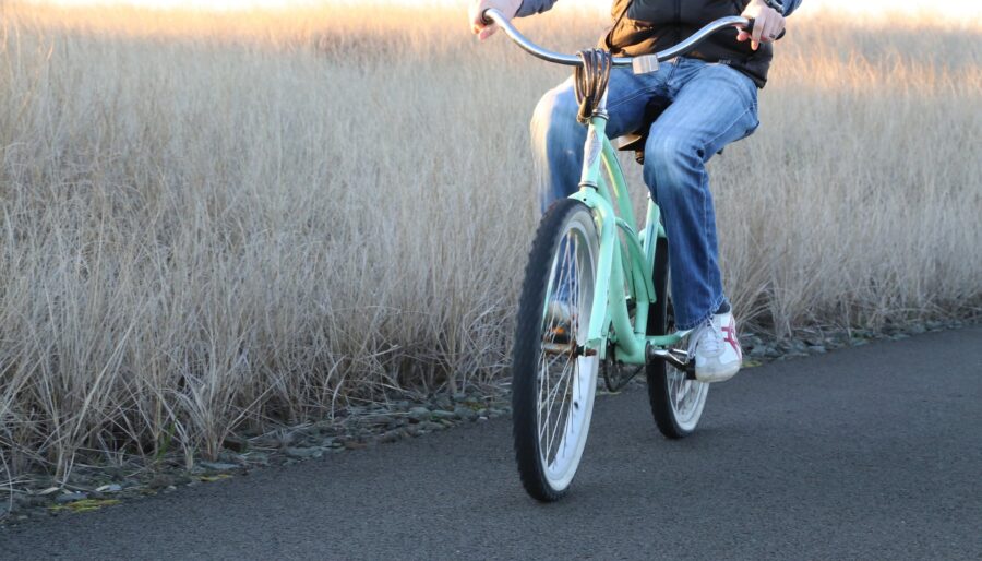 A person wearing a black jacket and blue jeans rides a light green bicycle on a paved path next to a field of tall, beige grass. The image showcases the person from the neck down.