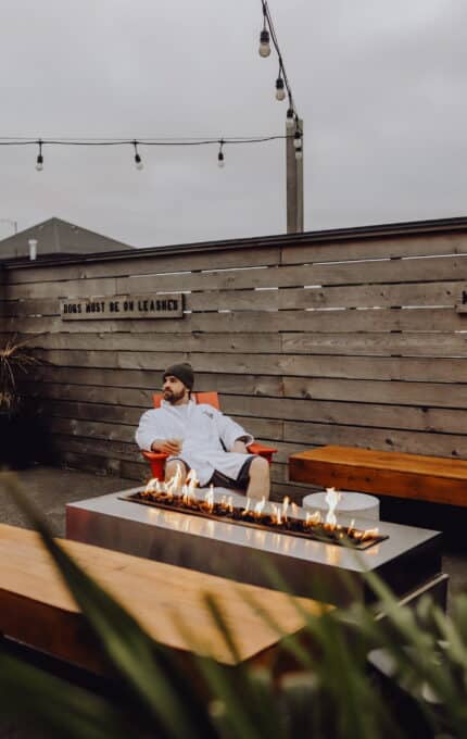 A person wearing a beanie and a robe sits in a red chair beside a modern outdoor firepit. The surroundings feature wooden walls and benches, string lights, and two signs that state "DOGS MUST BE ON LEASHES" and "NO SMOKING".