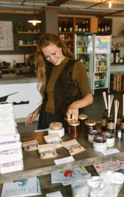 A person with long hair and a brown shirt is smiling and looking at various products on display in a shop. The products include jars, bottles with droppers, and packaged items. A refrigerator and shelves with bottles are visible in the background.