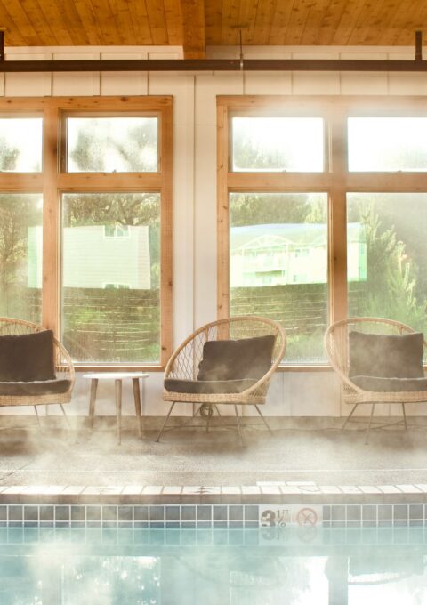 A serene indoor pool area with steam rising from the water. Several rattan chairs with dark cushions are lined up against large windows that let in natural light. The wooden ceiling adds a warm touch to the space.