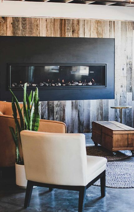 A modern lounge area features a wall-mounted fireplace on a wood-paneled wall, flanked by two brown leather chairs. A white chair and a wooden coffee table are centered on a round rug. A potted plant and side tables complete the setting.